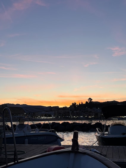 A sunset over a harbor with boats docked, a wooden pier, and buildings in the background. 4