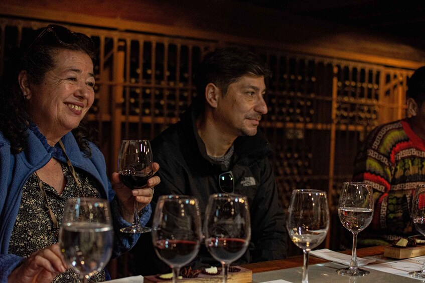 People seated at a table with wine glasses, a wooden backdrop with shelves, and a patterned garment. 3