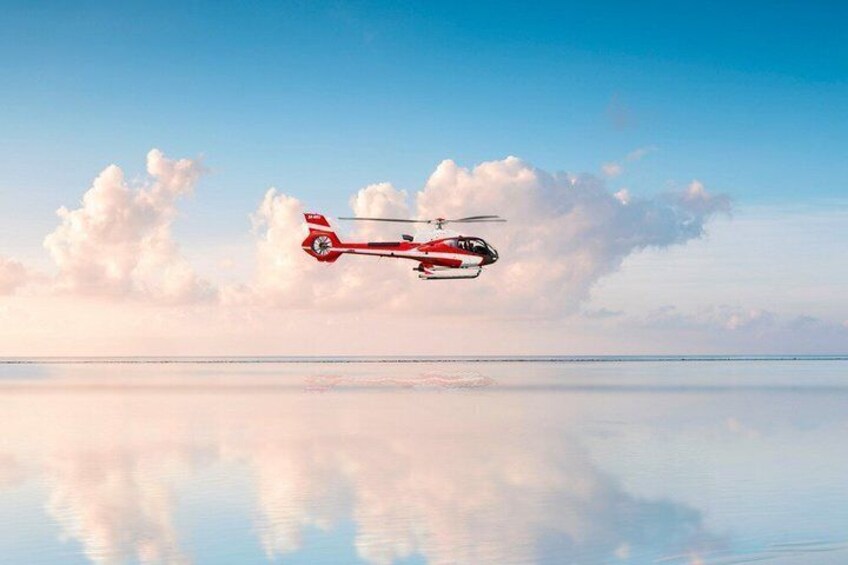 A red helicopter flying over a calm body of water with a clear reflection of the sky and clouds. 2
