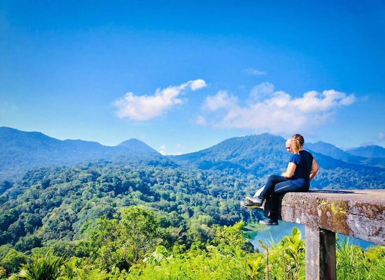 A person sitting on a concrete ledge overlooking a dense forest and mountains. 5