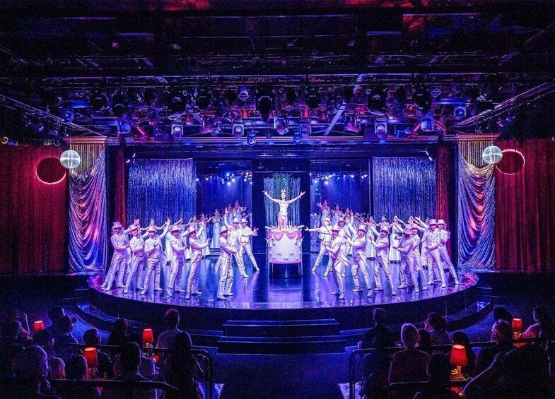 A stage with a group of performers in white costumes, a central figure on a platform, and a seated audience. 5