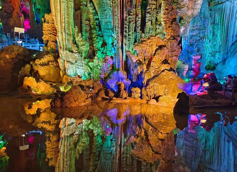 En grotta med färgstarka stalaktiter och stalagmiter, upplyst av artificiellt ljus, och en reflekterande vattenyta i förgrunden. 2