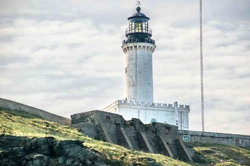 A lighthouse with a white tower and black top, situated on a grassy hill with a stone wall in the foreground. 2