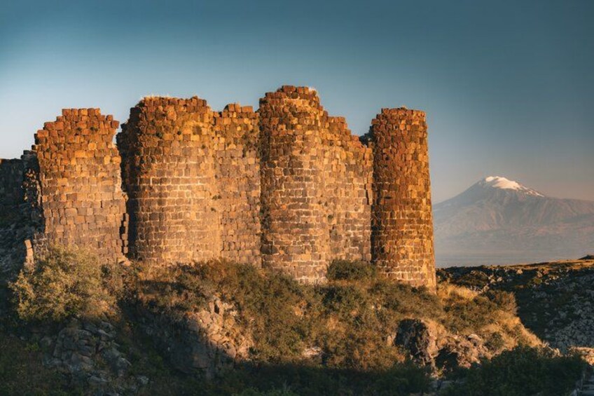 A historical stone castle with multiple towers, set against a mountainous backdrop with a snow-capped peak. 2