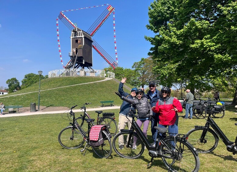 A group of people posing in front of a windmill, with bicycles and a park bench in the foreground. 5