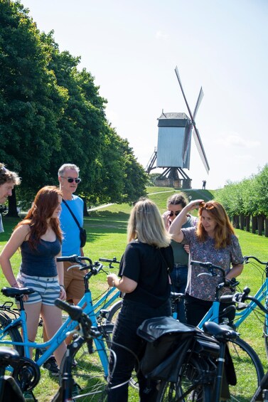 A group of people standing around bicycles in a park with a windmill in the background. 1