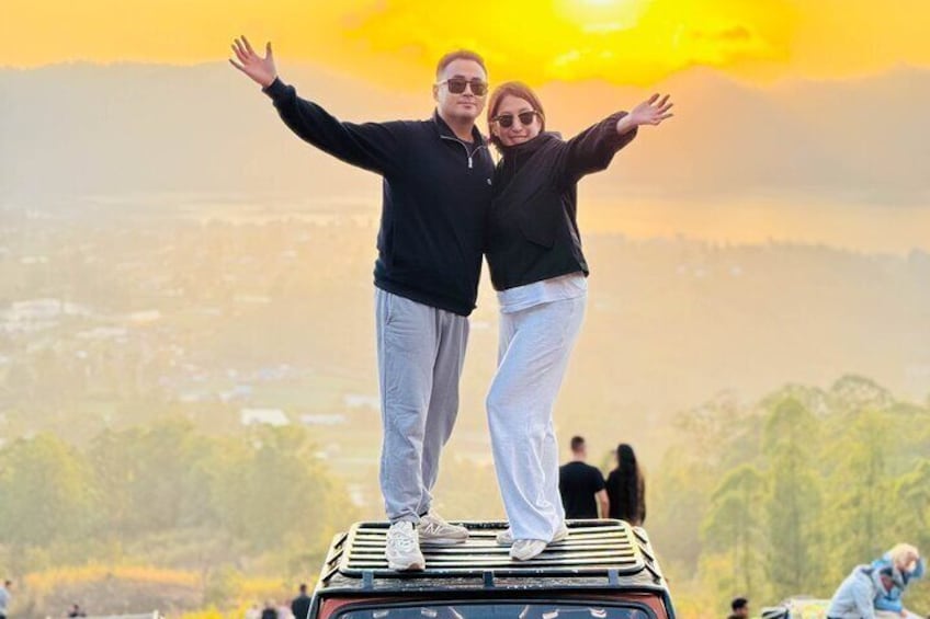 Two people standing on a vehicle with arms raised, overlooking a landscape with trees and a distant cityscape. 1