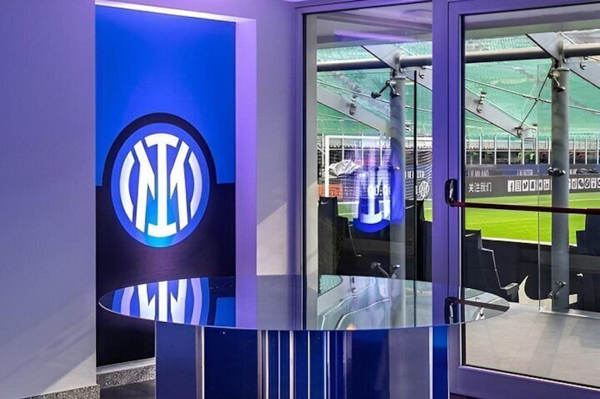 A modern indoor sports facility with a prominent logo on the wall and a glass door leading to a sports field. 1