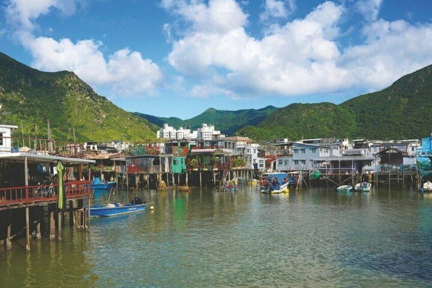 A coastal village with buildings on stilts, boats docked in the water, and green hills in the background. 1