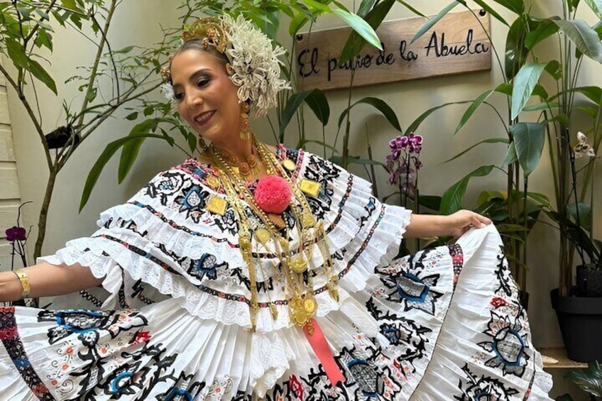 A person in a traditional embroidered dress with a floral headpiece, standing in a room with plants and a sign that reads 'El paƱo de la Abuela.' 1