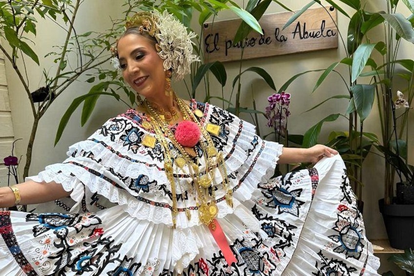 A person in a traditional embroidered dress with a floral headpiece, standing in front of a sign that reads 'El paƱo de la Abuela.' 1