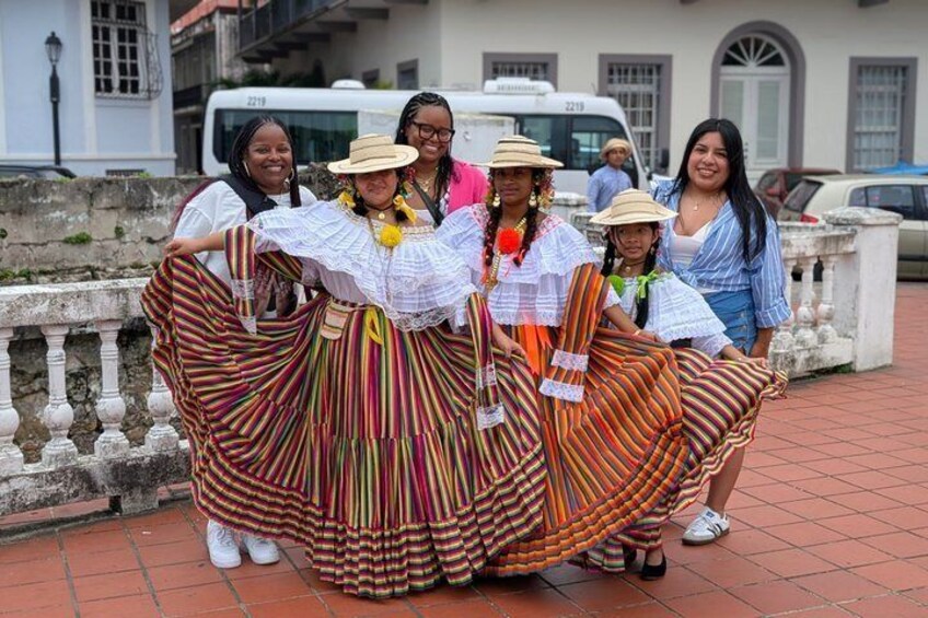 A group of people in traditional, colorful dresses with wide skirts, standing on a tiled pavement with a building and a white vehicle in the background. 4