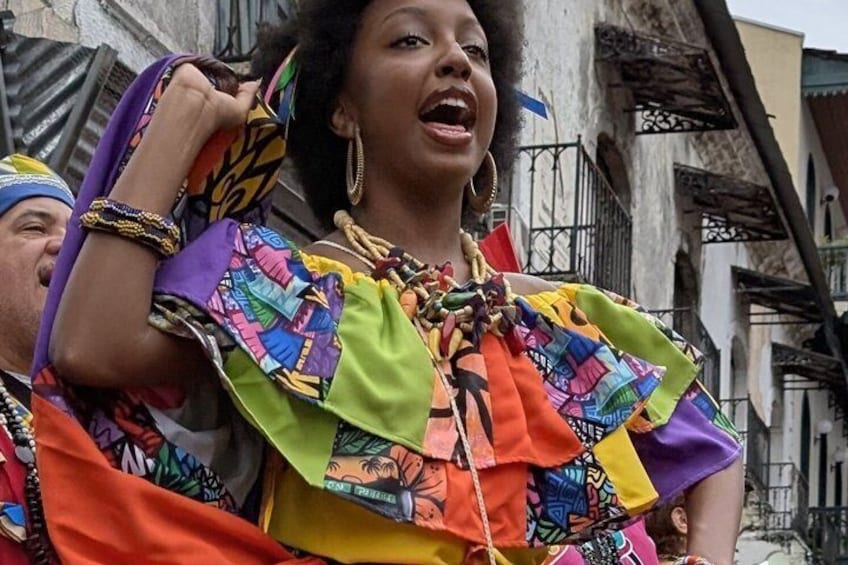 A person in a colorful, patterned outfit with ruffles, standing in an urban setting with buildings in the background. 2