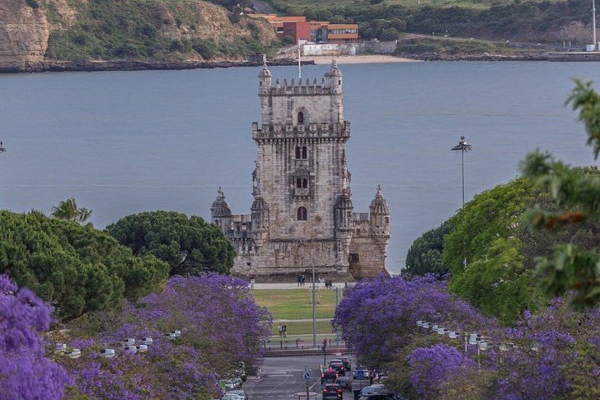 A historic tower by a body of water, surrounded by trees and purple flowers. 1