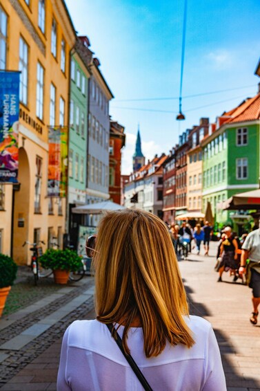A person with shoulder-length hair walking on a cobblestone street lined with colorful buildings. 3