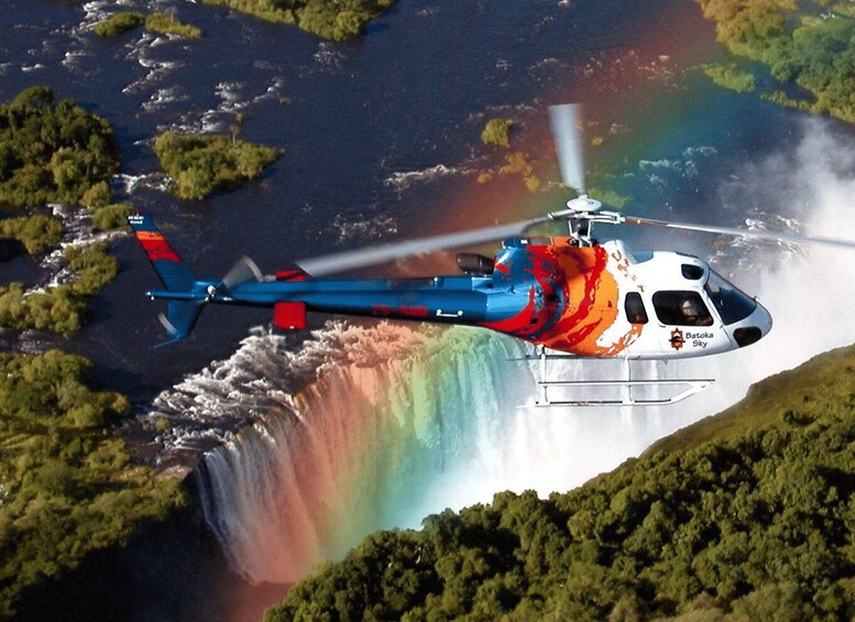 Helikopteri lentää vesiputouksen yläpuolella, ja taivaalla näkyy sateenkaari. 2
