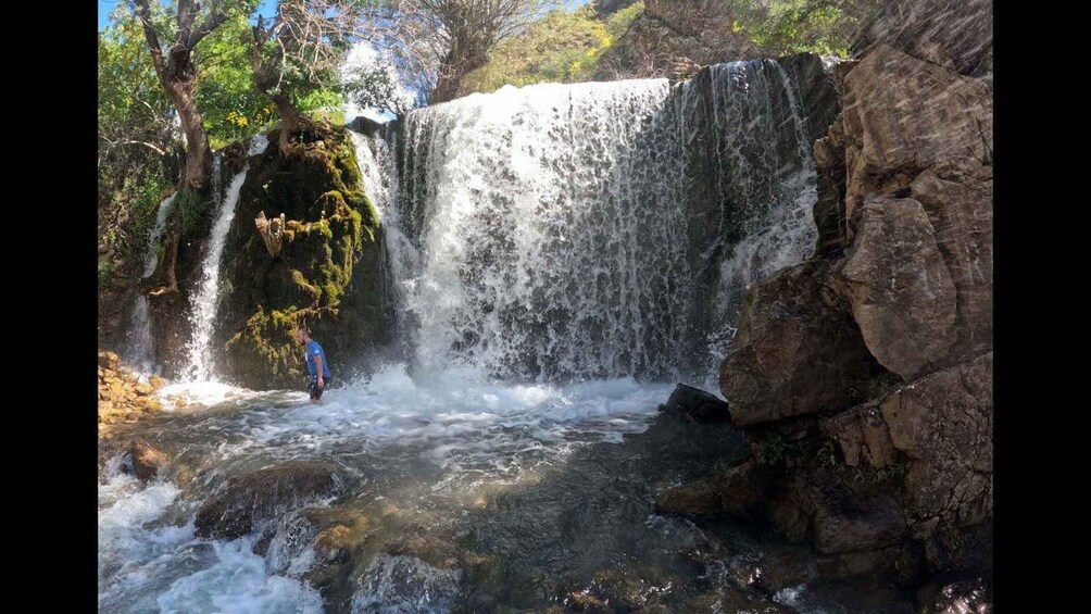 A waterfall cascading over rocks, a person standing in the water, and lush greenery surrounding the scene. 3