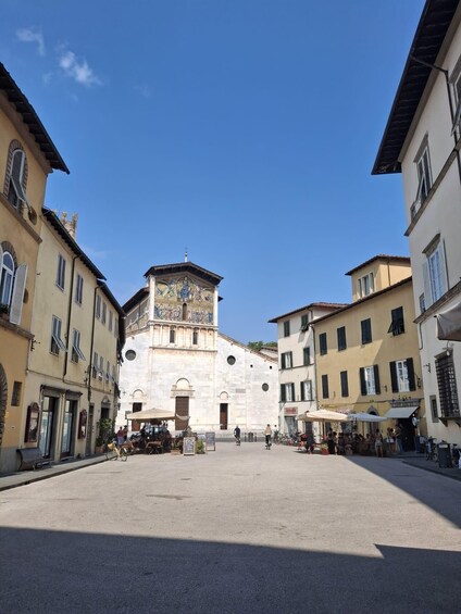 A square with a historic building featuring a clock tower and frescoes, surrounded by traditional buildings with shutters. 2