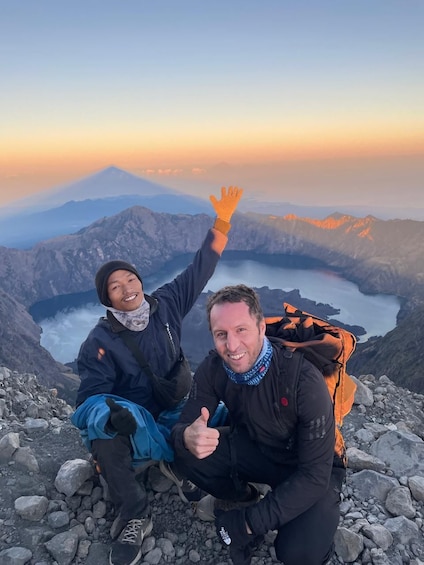 Two people sitting on a rocky mountain peak with a lake and a mountain in the background. 2