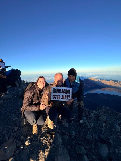 Three people on a mountain peak with a sign reading 'Rinjani 3726 MDPL'. 3