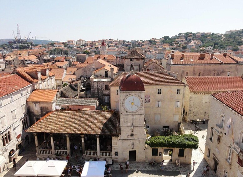 A historic town square with a prominent clock tower, terracotta-roofed buildings, and a bustling pedestrian area. 1