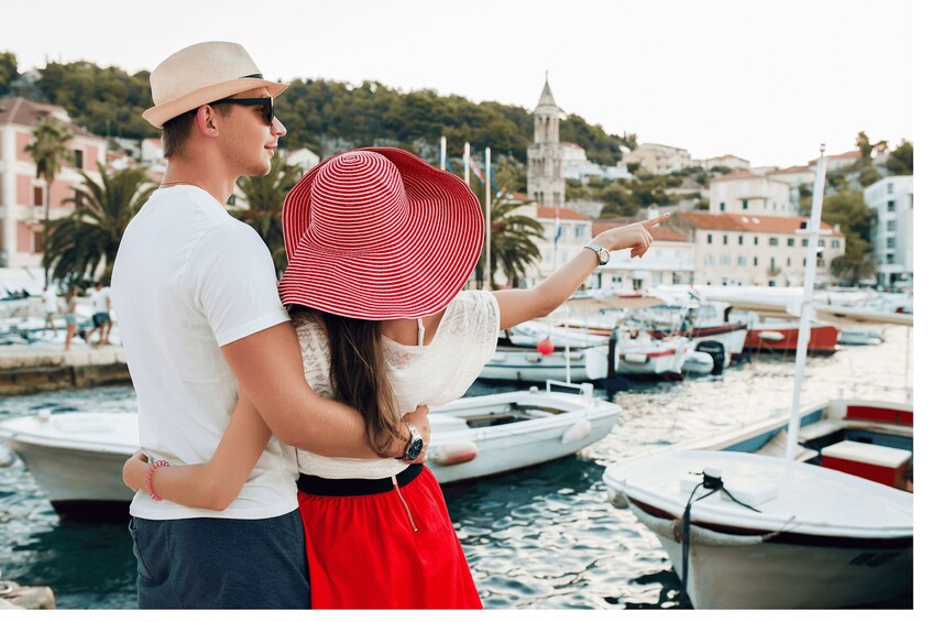 A couple standing by a marina with boats, a tower, and buildings in the background. 2