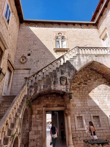 A stone building with a staircase leading to an entrance, a window with a decorative frame, and a clear blue sky above. 5