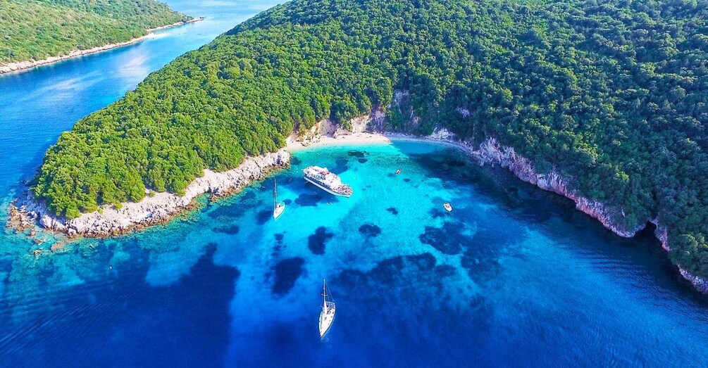 An aerial view of a turquoise bay surrounded by dense green forest, with a cruise ship docked and a sailboat anchored nearby. 1