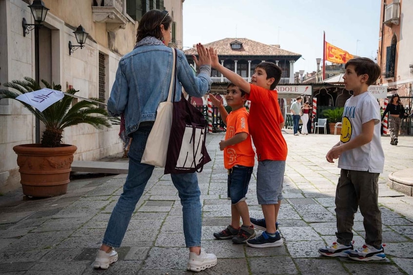 A woman in a denim jacket and white shoes walking with two children, one in an orange shirt and the other in a white shirt with a smiley face, on a cobblestone street with buildings and flags in the background. 2
