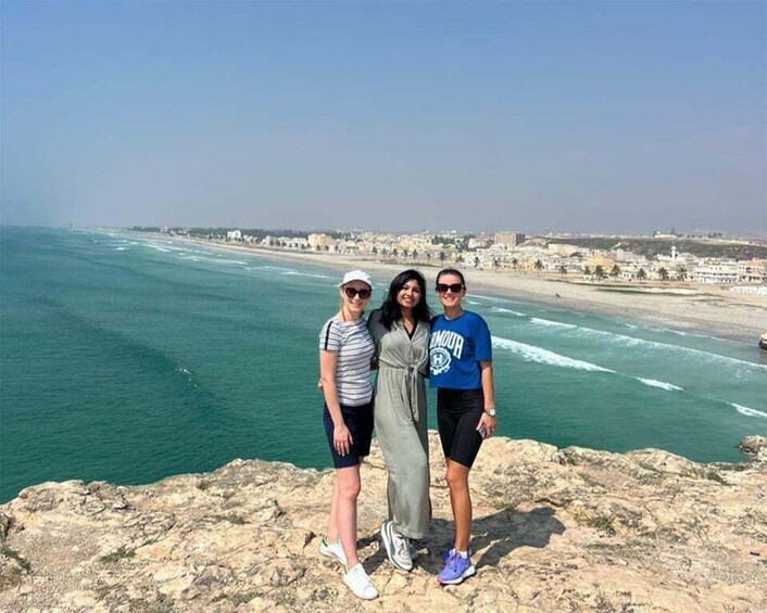 Tre persone in piedi su una sporgenza rocciosa, con una città costiera e una spiaggia sullo sfondo. 5