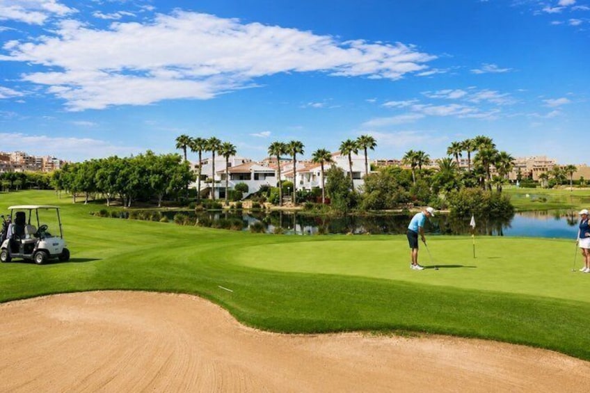 A golf course with a sand bunker, a water hazard, and a clubhouse in the background. 1