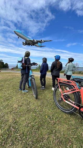 A group of people with bicycles and helmets, a blue bicycle, and a red bicycle, standing on grass with a plane in the sky. 2