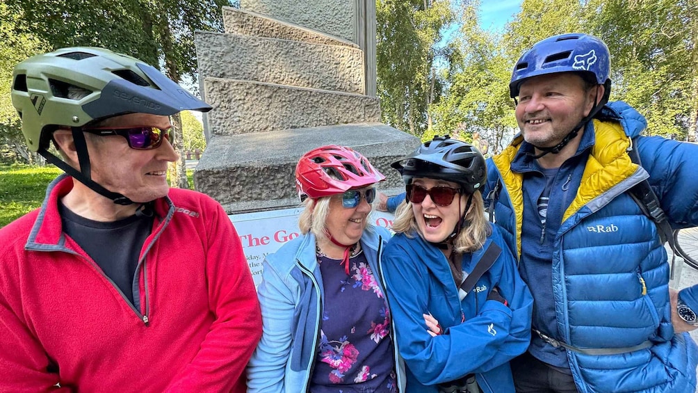 Three people wearing helmets and jackets, standing in front of a stone monument with a sign. 5