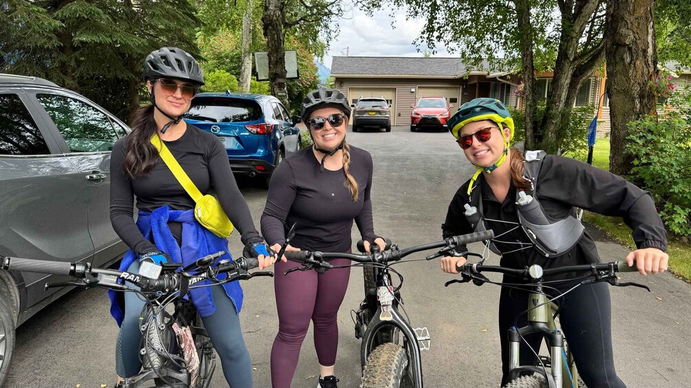 Three cyclists on a paved path with cars parked on the side, surrounded by trees and a building in the background. 4