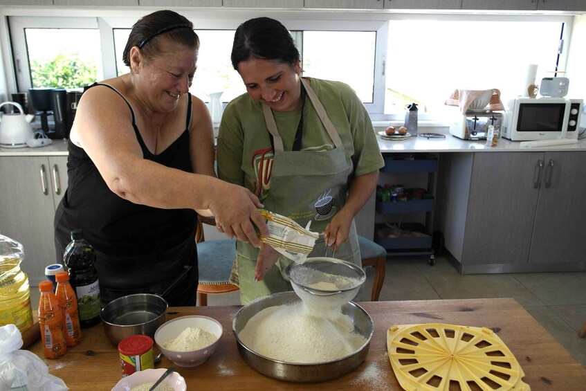 Two people in a kitchen preparing food, with ingredients and kitchen appliances visible. 1