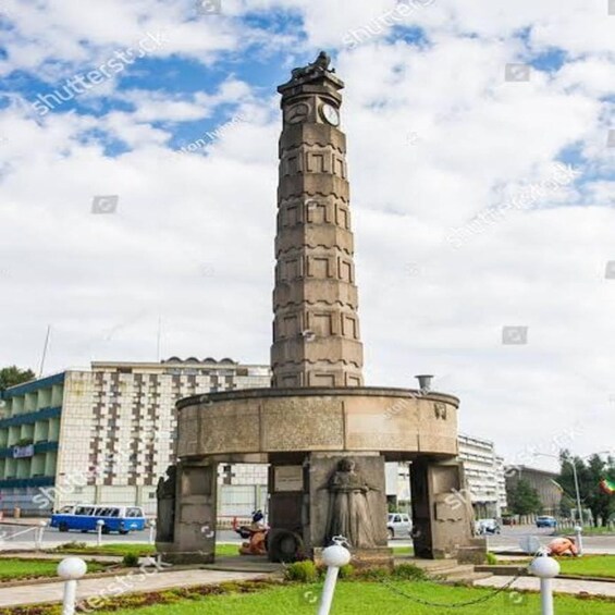 A tall stone monument with a clock, surrounded by a circular base and a statue, set in a grassy area with buildings in the background. 1
