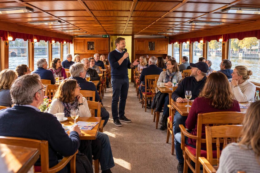 A boat with people seated at tables, a speaker addressing the audience, and a river view through large windows. 2
