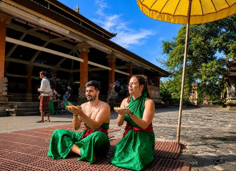 Two people in green traditional attire sitting on a patterned mat in front of a temple with a yellow umbrella. 5