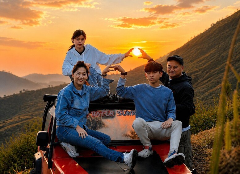 Four people posing with a red vehicle labeled 'BATUR SUNR' in a mountainous landscape during sunset. 1