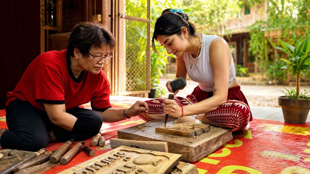 Two people engaged in wood carving on a red mat outdoors. 1