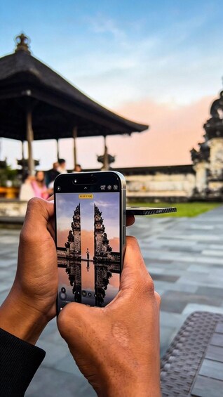 A person holding a smartphone, capturing a reflection of a temple's entrance in a body of water. 2