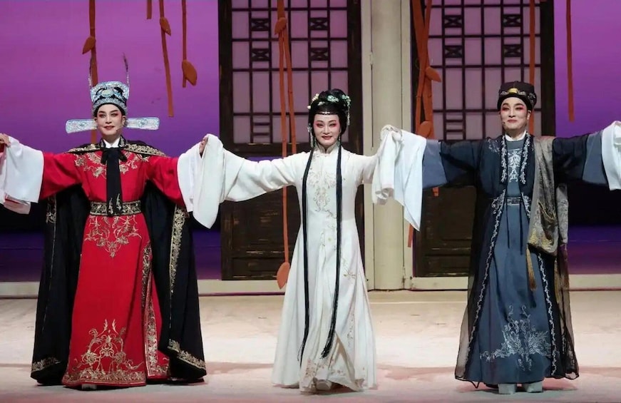 Three individuals in traditional costumes with outstretched arms, standing on a stage with a purple backdrop. 2