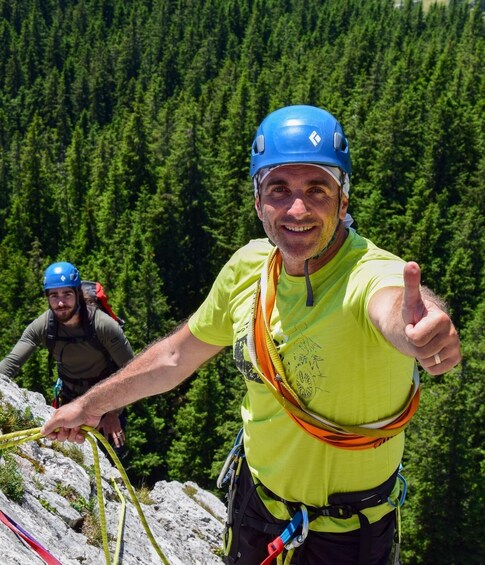 A person giving a thumbs up, wearing a blue helmet and a yellow shirt, with a forested mountain background. 1