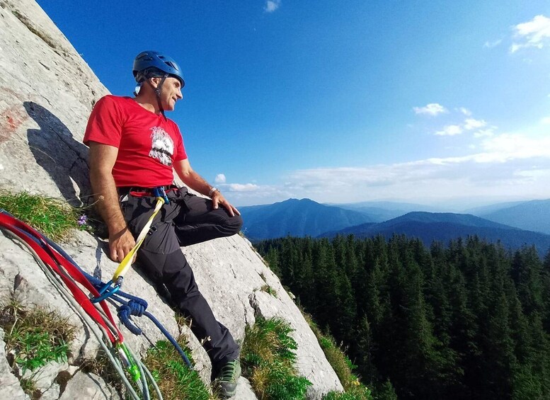 A person sitting on a rock with climbing gear, overlooking a forested mountain range. 4