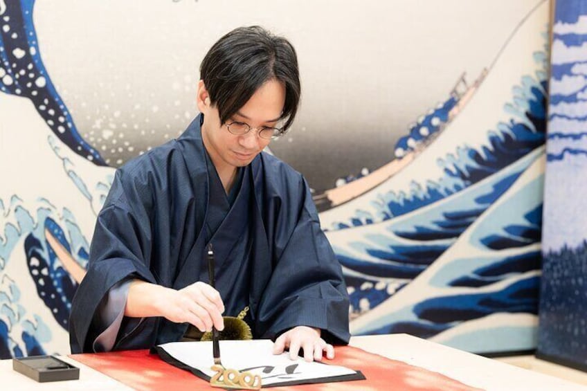 A person in traditional attire sitting at a table with a tablet and a pen, in front of a Japanese wave painting. 1