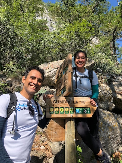 Two people standing next to a wooden signpost with hiking symbols and a mountain trail name. 2