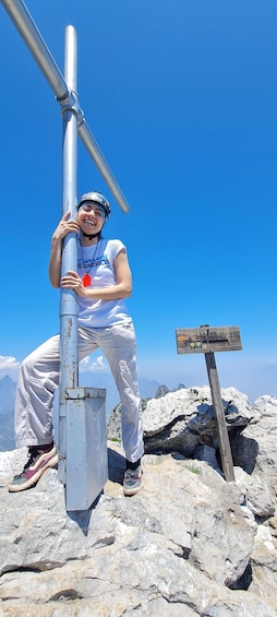 A person standing on a rocky mountain peak with a metal structure and a wooden sign in the background. 3