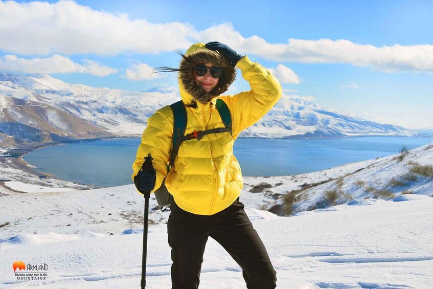 Una persona con una chaqueta amarilla y pantalones negros de pie en una montaña nevada, con un cielo azul despejado y un lago al fondo. 1