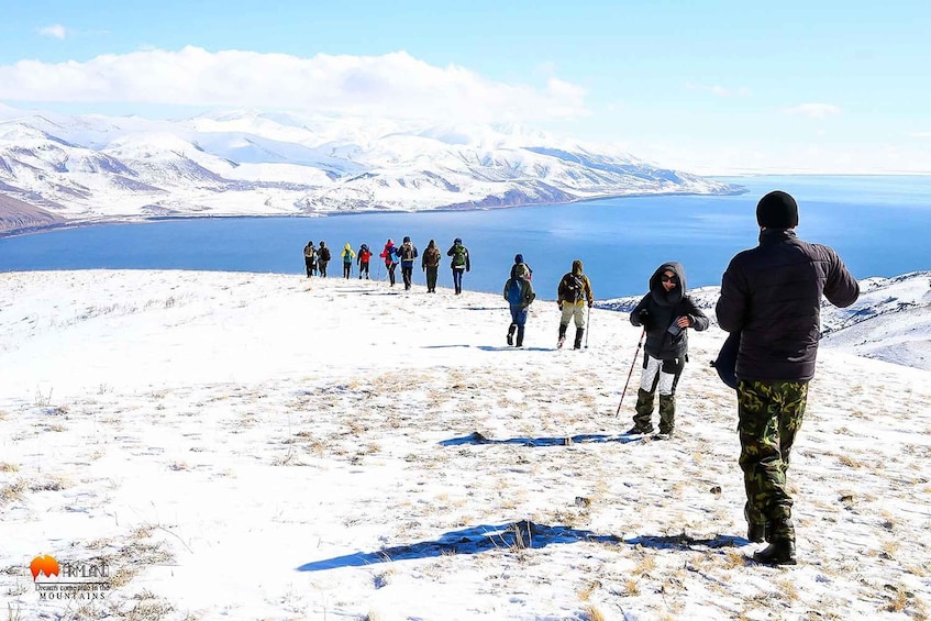 Un grupo de excursionistas en un sendero nevado de montaña, con un lago y montañas cubiertas de nieve al fondo. 2