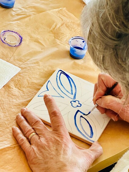 An elderly person painting on a small canvas with blue paint, surrounded by paint containers and a tissue. 1